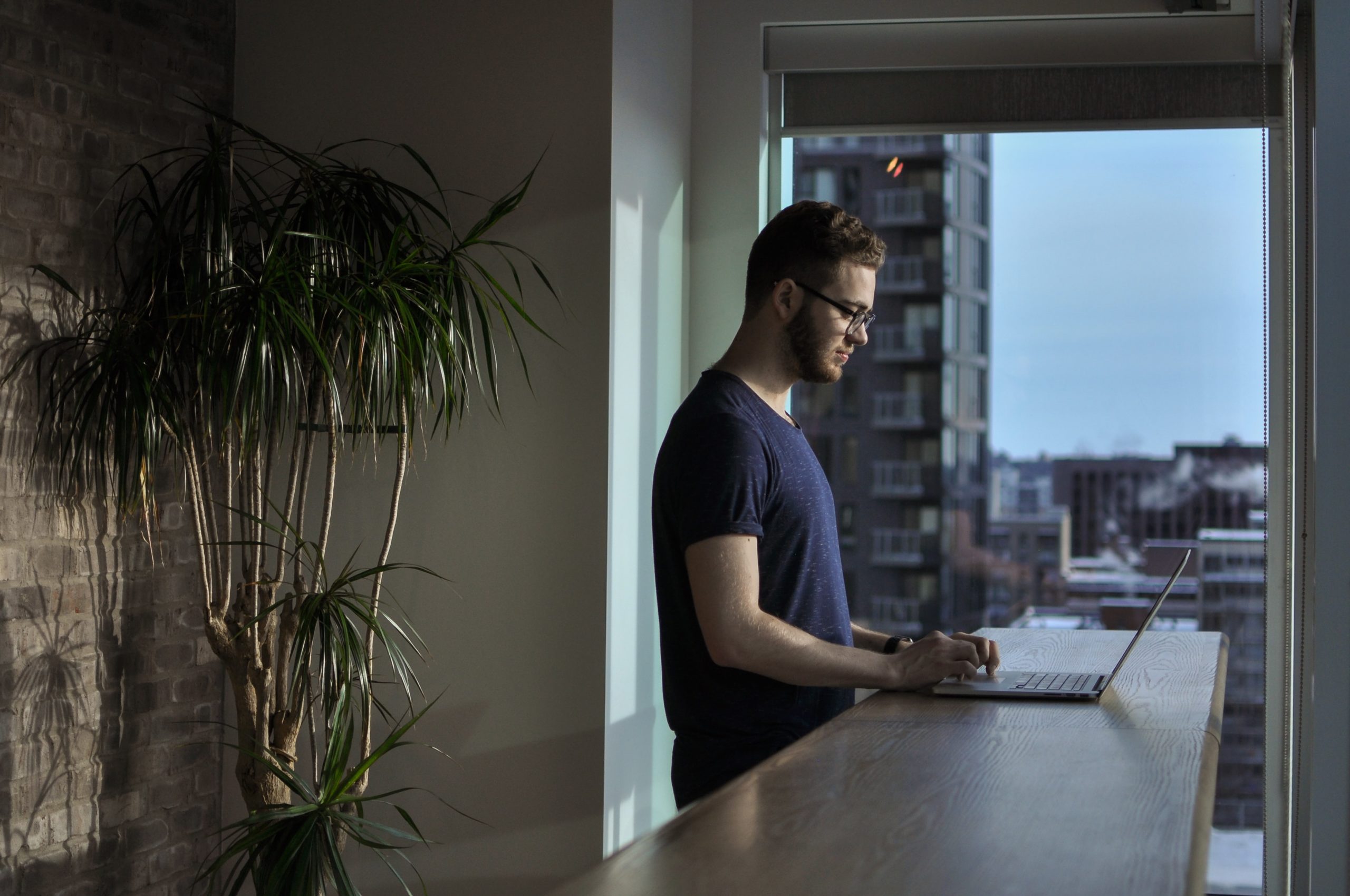 Article Cover: Photo of a man standing working on a laptop in front of a large window